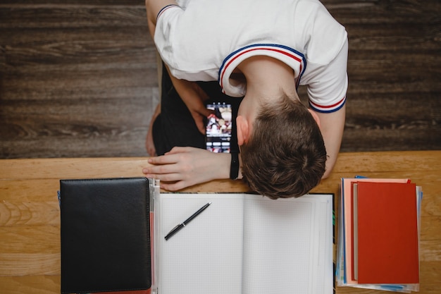 Foto un adolescente en casa para libros de texto en lugar de lecciones mira un teléfono inteligente en secreto debajo de la mesa