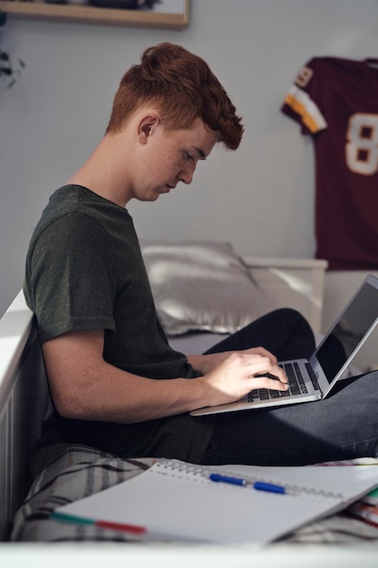 Foto adolescente caucasiano sentado na cama e aprendendo usando laptop
