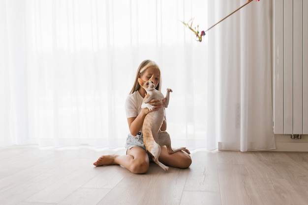 Una adolescente caucásica juega con un gato Cornish Rex en casa en un día soleado, lo abraza y se ríe mientras se sienta en el suelo en la sala de estar