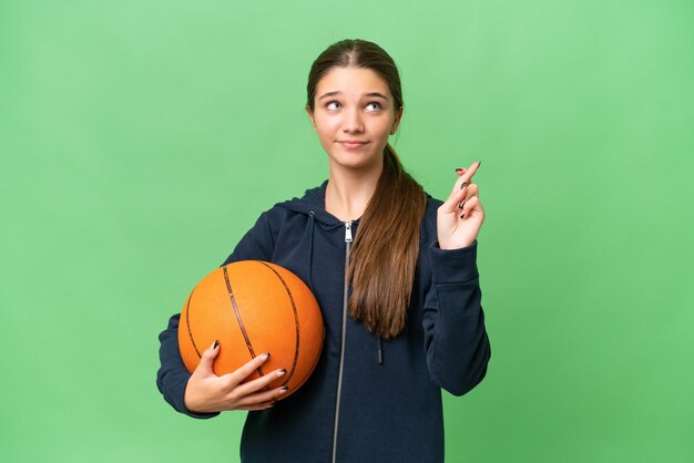 Adolescente caucásica jugando baloncesto sobre un fondo aislado con los dedos cruzados y deseando lo mejor