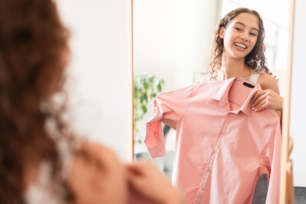 Una adolescente caucásica sonriente probándose una camisa cerca del espejo interior