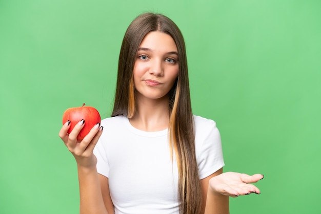 Adolescente caucásica sosteniendo una manzana sobre un fondo aislado haciendo gestos de duda mientras levanta los hombros