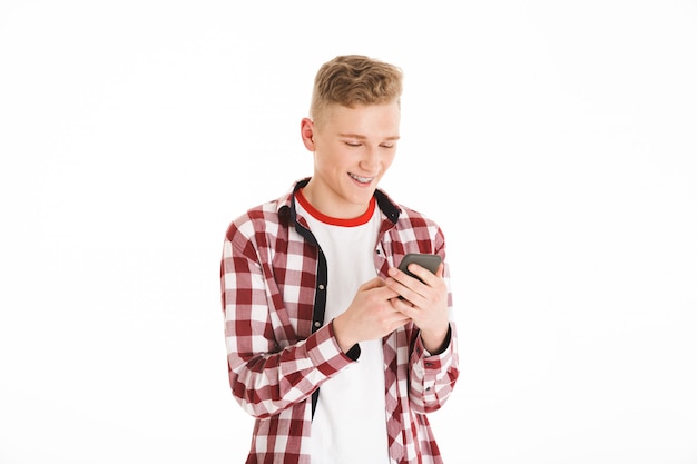 Adolescente caucásico en camiseta casual con brackets sosteniendo el teléfono celular y mirando la pantalla mientras navega o escribe, aislado sobre la pared blanca