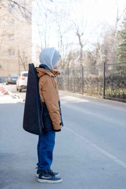 Un adolescente con chaqueta y sombrero camina solo con una guitarra en un estuche Niño músico perdido en la ciudad