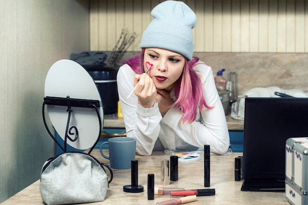 Foto adolescente chica con cabello rosado hace maquillaje brillante en casa frente al espejo.