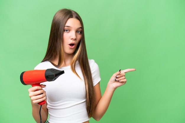 Adolescente chica caucásica sosteniendo un secador de pelo sobre un fondo aislado sorprendido y apuntando hacia el lado