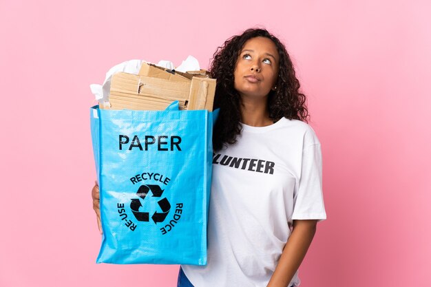 Adolescente chica cubana sosteniendo una bolsa de reciclaje llena de papel para reciclar aislado sobre fondo rosa y mirando hacia arriba