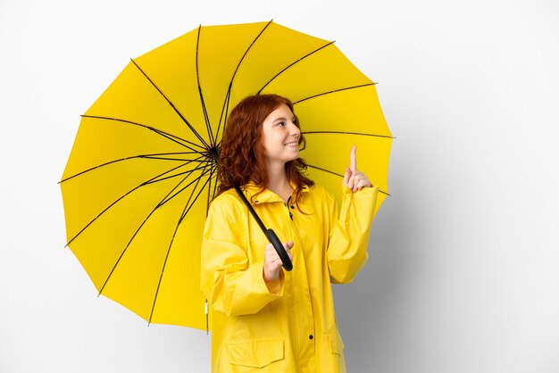 Adolescente chica pelirroja abrigo impermeable y paraguas aislado sobre fondo blanco apuntando hacia una gran idea