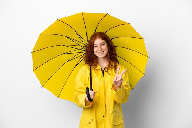 Adolescente chica pelirroja abrigo impermeable y paraguas aislado sobre fondo blanco sonriendo y mostrando el signo de la victoria