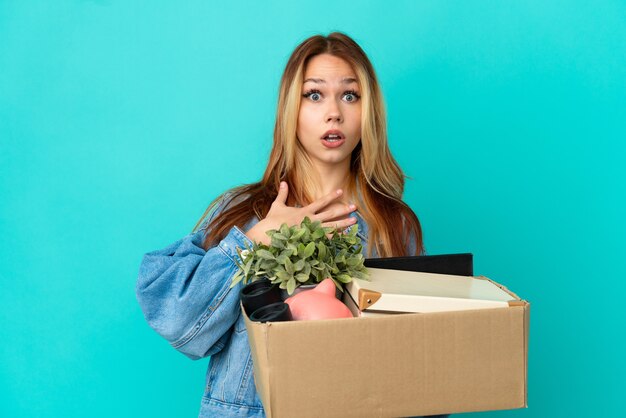 Adolescente chica rubia haciendo un movimiento mientras recoge una caja llena de cosas sorprendido y conmocionado mientras mira a la derecha