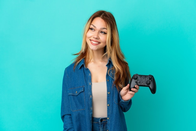 Adolescente chica rubia jugando con un controlador de videojuegos sobre pared aislada mirando hacia el lado y sonriendo