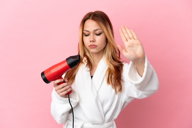 Adolescente chica rubia sosteniendo un secador de pelo sobre fondo rosa aislado haciendo gesto de parada y decepcionado