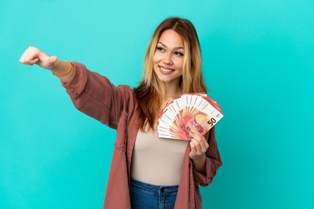 Adolescente chica rubia teniendo un montón de euros sobre fondo azul aislado dando un gesto de pulgar hacia arriba