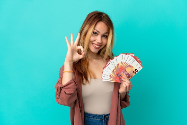 Adolescente chica rubia teniendo un montón de euros sobre fondo azul aislado mostrando un signo de ok con los dedos