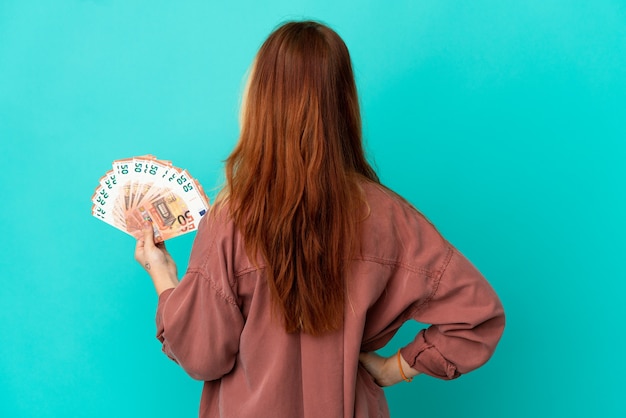 Adolescente chica rubia teniendo un montón de euros sobre fondo azul aislado en posición trasera