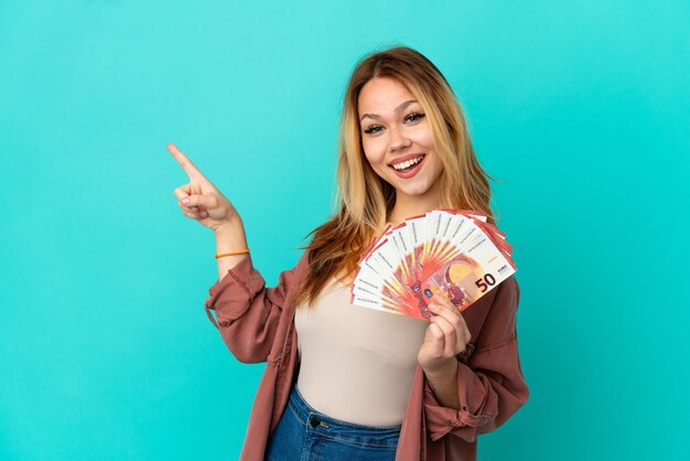 Foto adolescente chica rubia teniendo un montón de euros sobre fondo azul aislado que señala el dedo hacia el lado y presenta un producto