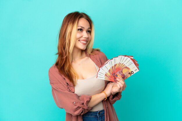 Adolescente chica rubia tomando un montón de euros sobre fondo azul aislado feliz y sonriente