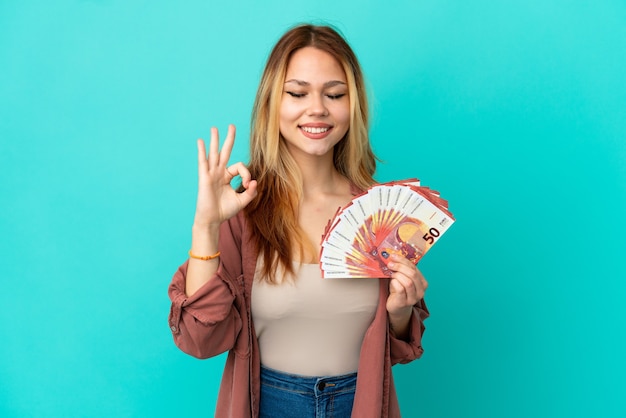 Adolescente chica rubia tomando un montón de euros sobre fondo azul aislado en pose zen