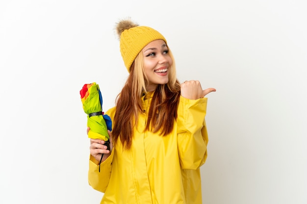 Adolescente chica rubia vistiendo un abrigo impermeable sobre fondo blanco aislado apuntando hacia el lado para presentar un producto