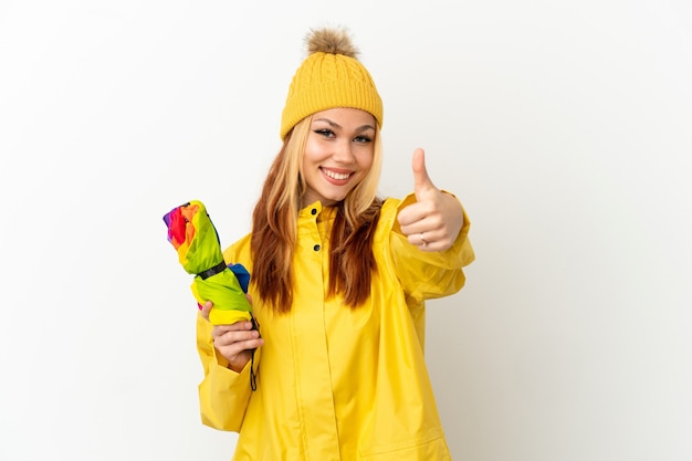 Adolescente chica rubia vistiendo un abrigo impermeable sobre fondo blanco aislado con los pulgares hacia arriba porque algo bueno ha sucedido