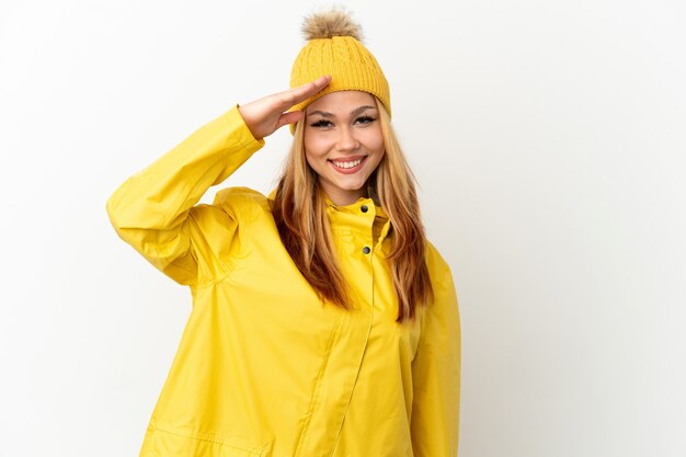 Adolescente chica rubia vistiendo un abrigo impermeable sobre fondo blanco aislado saludando con la mano con expresión feliz