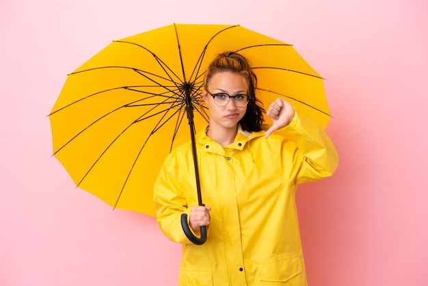 Adolescente chica rusa con abrigo impermeable y paraguas aislado sobre fondo rosa mostrando el pulgar hacia abajo con expresión negativa