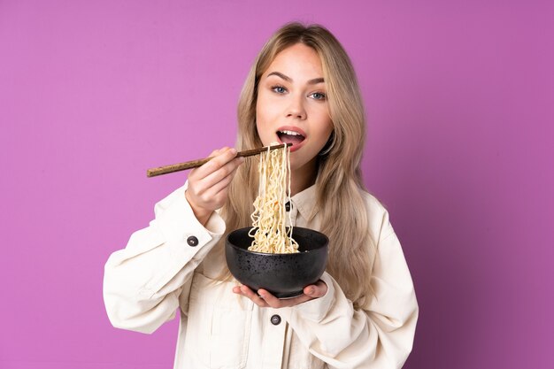 Adolescente chica rusa aislada en la pared púrpura sosteniendo un plato de fideos con palillos y comiéndolo