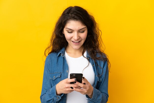 Adolescente chica rusa aislada sobre fondo amarillo enviando un mensaje o correo electrónico con el móvil