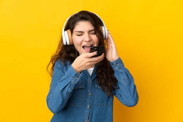 Adolescente chica rusa aislada sobre fondo amarillo escuchando música con un móvil y cantando