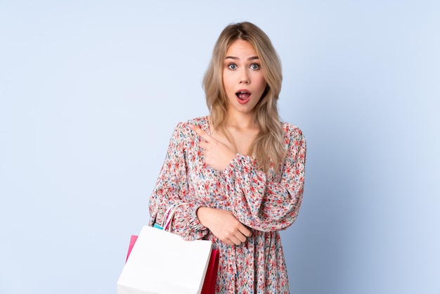 Adolescente chica rusa con bolsa de compras aislado en la pared azul sorprendido y apuntando hacia el lado