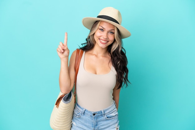 Adolescente chica rusa con pamel y bolsa de playa aislado sobre fondo azul mostrando y levantando un dedo en señal de lo mejor