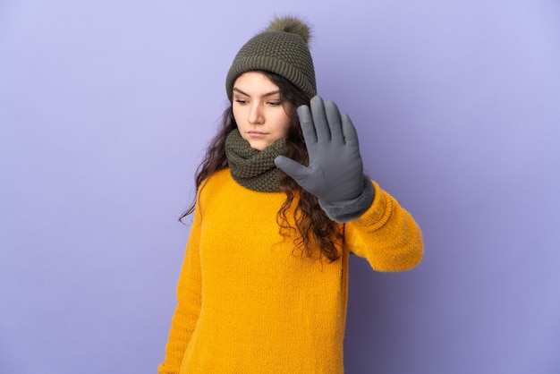 Adolescente chica rusa con sombrero de invierno aislado sobre fondo púrpura haciendo gesto de parada y decepcionado