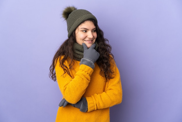 Adolescente chica rusa con sombrero de invierno aislado sobre fondo púrpura mirando hacia el lado y sonriendo