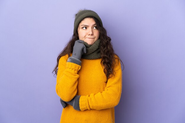 Adolescente chica rusa con sombrero de invierno aislado en superficie púrpura con dudas y pensamiento