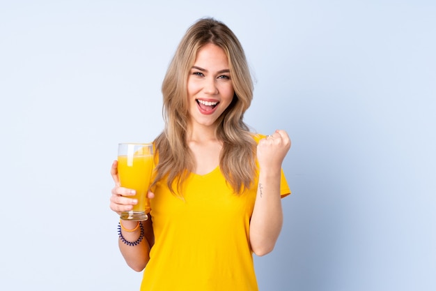 Adolescente chica rusa sosteniendo un jugo de naranja aislado sobre fondo azul celebrando una victoria