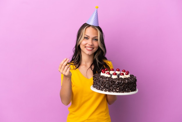 Adolescente chica rusa sosteniendo pastel de cumpleaños aislado sobre fondo púrpura haciendo gesto de dinero