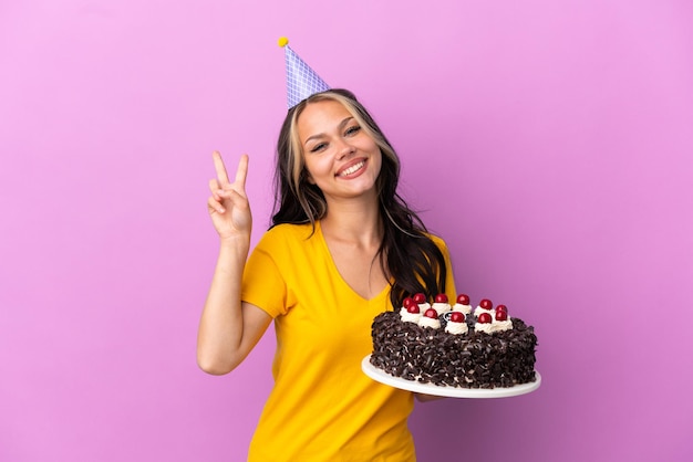 Adolescente chica rusa sosteniendo pastel de cumpleaños aislado sobre fondo púrpura sonriendo y mostrando el signo de la victoria