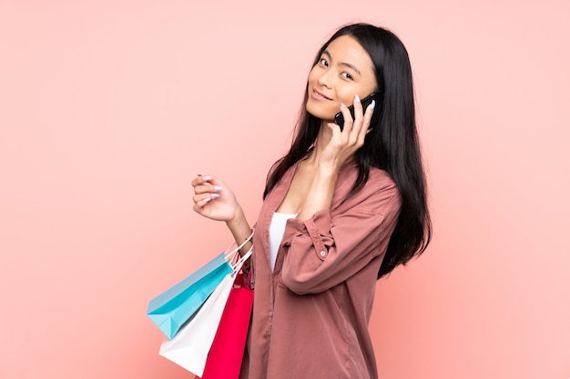 Adolescente china en la pared rosa sosteniendo bolsas de compras y llamando a un amigo con su teléfono celular
