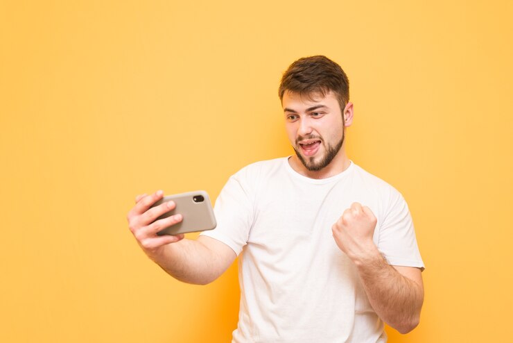 Foto adolescente com barba jogando jogos para celular no smartphone, olhando para a tela e se alegrando