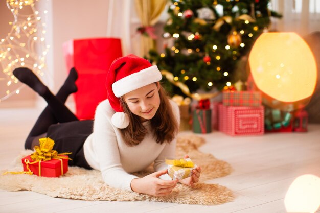 Adolescente com chapéu de papai noel abrindo seu presente de natal deitado no tapete