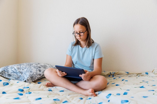 Adolescente com óculos jogando um tablet em uma cama no quarto