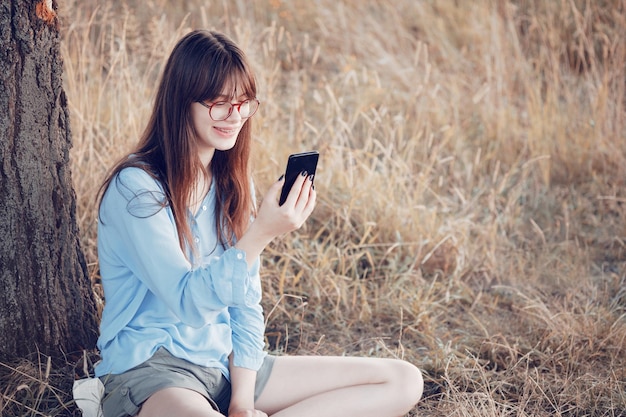Adolescente com óculos senta-se perto de uma árvore no gramado e olha para o telefone