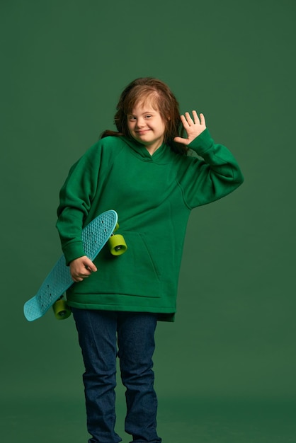 Adolescente com síndrome de Down vestindo um capuz aconchegante de pé com skate contra um estúdio verde