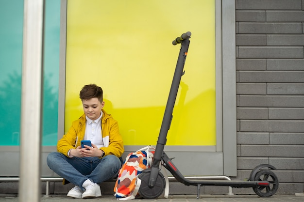 Adolescente com telefone celular sentado ao lado de sua scooter elétrica e mochila.
