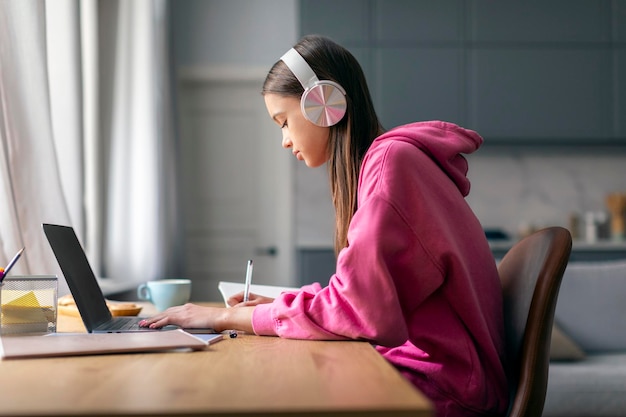 Adolescente concentrada con auriculares inalámbricos sentada en el escritorio en casa estudiando en la computadora portátil haciendo
