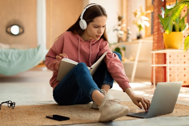 Una adolescente concentrada leyendo un libro frente a una laptop preparándose para el examen sentada en el suelo en casa