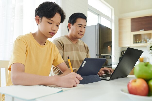 Adolescente concentrado haciendo los deberes en la mesa de la cocina cuando está sentado junto a su padre trabajando en la computadora portátil