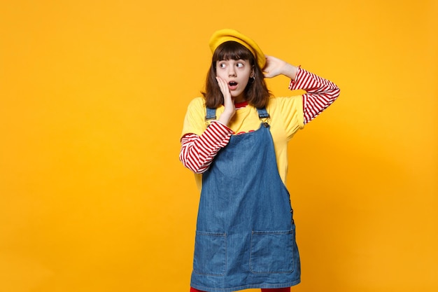 Adolescente confuso chocado na boina francesa, vestido jeans mantendo a boca aberta, colocando a mão na bochecha, olhando de lado isolado no fundo da parede amarela. emoções sinceras de pessoas, conceito de estilo de vida.