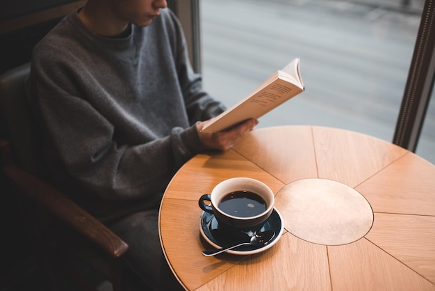 Adolescente de 17 a 18 anos lendo livro de papel e bebendo café preto na mesa de madeira no café close-up. Jovem milenar estuda e trabalha com uma xícara de café expresso. Local de trabalho