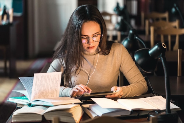 Foto adolescente de cabelos compridos pensativo estudando à mesa na biblioteca usando tablet e livros